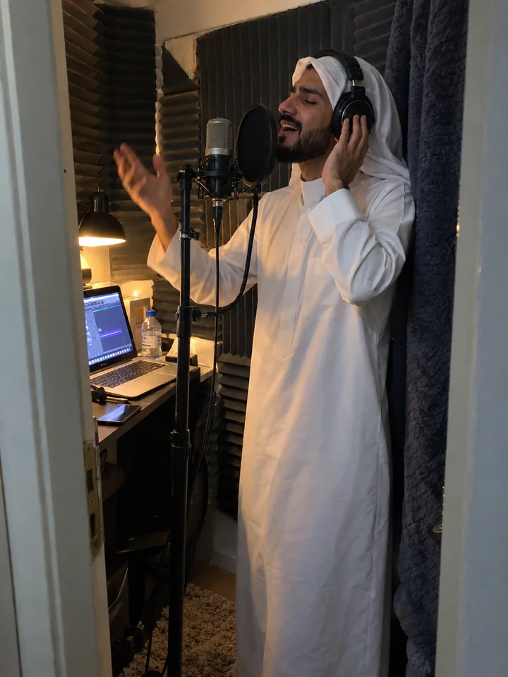 A young Saudi man singing into a microphone in a home recording booth, a phone photo